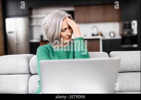 Donna d'affari matura esausta freelancer femminile seduta sul divano, toccando templi massaggianti che soffrono di grave mal di testa o emicrania, vecchia donna etnica stanca sopraffatta dal lavoro al computer Foto Stock