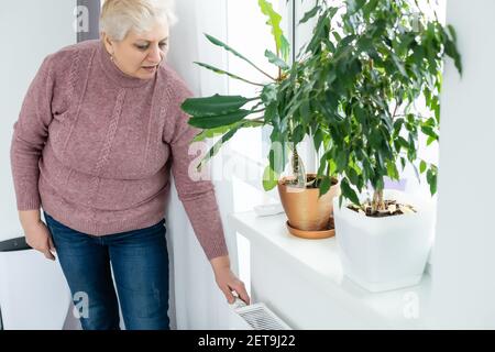 Donna anziana che cerca di mantenere caldo riscaldando le mani il radiatore di riscaldamento in inverno Foto Stock