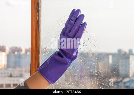 Una mano della donna in un guanto blu spugne una finestra polverosa con cornice in legno. Foto Stock