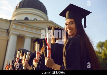 Donna laureata in piedi con un diploma in mano e guardando la telecamera indietro. Foto Stock