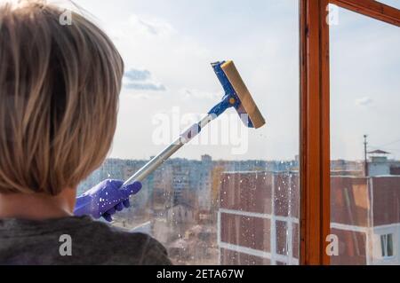 Una giovane donna irriconoscibile in guanti blu lava la finestra dall'esterno con un raschiatore speciale su un lungo maniglia Foto Stock