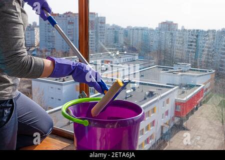 Una donna irriconoscibile che indossa guanti protettivi lava le finestre di un balcone con pannelli in legno con un mop speciale e una plastica secchio di età di pulizia Foto Stock