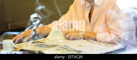 Una donna anziana in una camicia rossa a righe sta girando attraverso il giornale mentre sorseggiando il caffè la mattina. Stile di vita e persone, nessun volto visibile. Foto Stock