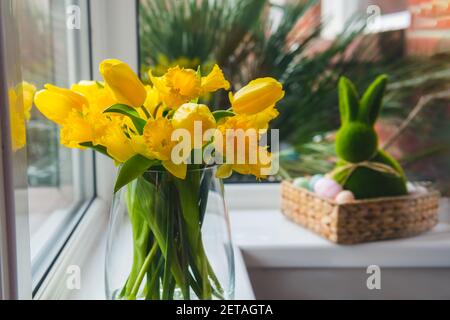 Bouquet di tulipani freschi di primavera e fiori di narcisi sul davanzale con coniglio coniglietto pasquale in cestino di paglia con uova colorate sullo sfondo. Felice Foto Stock