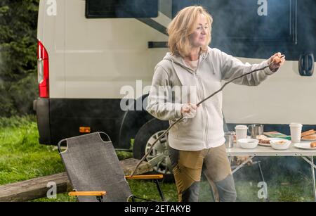 Donna caucasica nei suoi 40 avendo divertimento su un campeggio. Preparare la salsiccia polacca sul suo bastone da cucina fatto da sé. Moderno Motorhome di classe B in background. Foto Stock