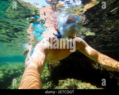 Vista subacquea di un giovane subacqueo che nuota nella mare turchese sotto la superficie con maschera da snorkeling per l'estate vacanze mentre si prende un selfie wi Foto Stock