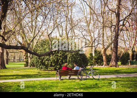 BUDAPEST, UNGHERIA - 04 APRILE 2019: La coppia è seduta su una panchina di legno sotto un albero nella bella giornata di primavera soleggiata sull'isola di Margeret a Budapest, Hun Foto Stock