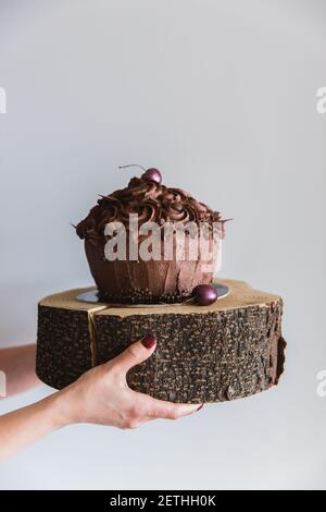 Donna che tiene una torta di cioccolato decorata con ciliegia su un piatto di legno, primo piano. Deliziosa panetteria e fornire alte calorie Foto Stock