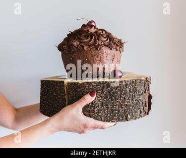 Donna che tiene una torta di cioccolato decorata con ciliegia su un piatto di legno, primo piano. Deliziosa panetteria e fornire alte calorie Foto Stock