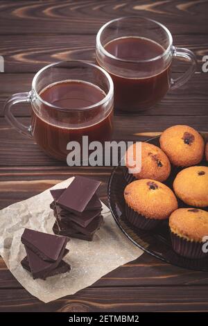 Pezzi di cioccolato fondente, muffin e due tazze con bevanda calda al cacao su uno sfondo di tavole di legno marroni. Il concetto di colazione romantica. Bevande Foto Stock