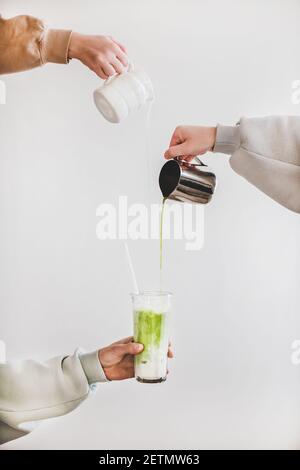 Le mani femminili versano il tè giapponese matcha e il latte dalla caraffa in un bicchiere per preparare il latte verde matcha per la colazione su uno sfondo chiaro della parete. Bevande al caffè e cucina giapponese Foto Stock