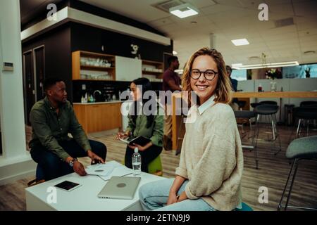 Donna caucasica sorridente prendendo pausa pranzo seduto in caffetteria mentre i colleghi ricoprono il lavoro la settimana seduti su comode sedie dentro sala ufficio Foto Stock