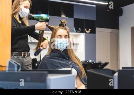 Master donna parrucchiere in una maschera medica asciuga i capelli della ragazza con un asciugacapelli e pettini dopo il lavaggio in un salone di bellezza. Covid-19 pandemia e. Foto Stock