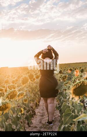 Una bella giovane ragazza con la schiena si girò a toccarla capelli e camminare attraverso un campo di girasoli in un tramonto di una giornata estiva Foto Stock