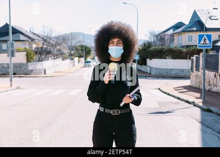 Giornalista televisiva femminile con capelli afro, indossando una maschera chirurgica per proteggersi da un virus, che riporta su una storia per strada Foto Stock