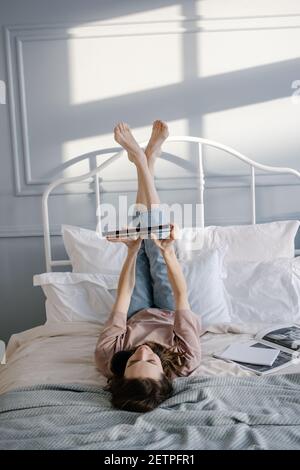 La donna creativa è sdraiata sul letto con il tablet, sorridente pensieroso, scrive un messaggio, chiama via video chat, legge le notizie. Lavoro da casa, freelance e. Foto Stock