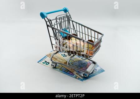 Carrello di acquisto del supermercato pieno di monete in euro sopra le banconote in euro. Concetto di costo di acquisto Foto Stock