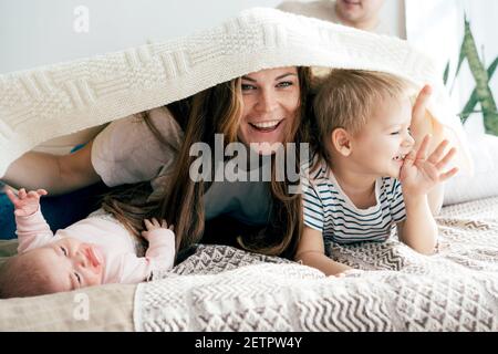 La mummia felice gioca con i bambini sul letto e copre i bambini e se stessa con una coperta. Divertimento per tutta la famiglia. Foto Stock