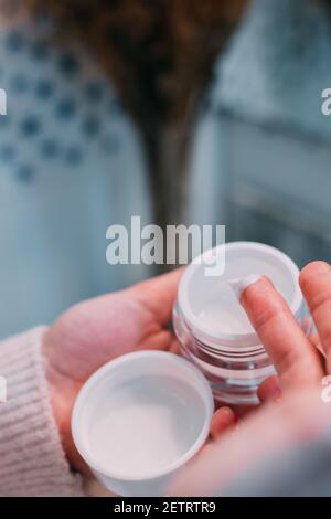 mano di una giovane donna che applica crema viso da un vaso di idratante in bagno nella sua routine di cura quotidiana. cura del corpo e concetto di bellezza Foto Stock