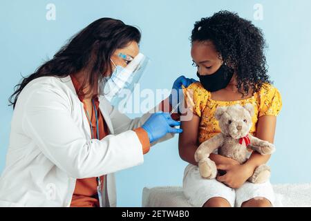 Medico femminile che indossa maschera protettiva, guanti e scudo iniettando il vaccino nel braccio della ragazza in maschera durante la pandemia. Piccola ragazza che la tiene il suo teddy Foto Stock