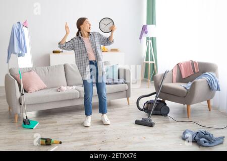 Scioccata giovane donna che tiene l'orologio, sentendosi terrorizzata, in piedi in disordinato stanza dopo la festa, essendo in ritardo per pulire l'appartamento Foto Stock