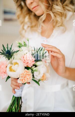 La sposa tiene in mano e tocca delicatamente un bouquet con peonie, rose, lisianto ed eringi Foto Stock