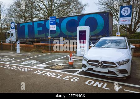 Mercedes carica auto in una stazione di ricarica elettrica presso un negozio Tesco nella città di Uckfield, Sussex, Regno Unito con un camion Tesco consegna in background. Foto Stock