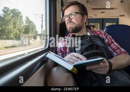 L'uomo caucasico serio guida un treno e guarda pensieroso fuori la finestra, in mani tiene un libro e una matita. Foto Stock