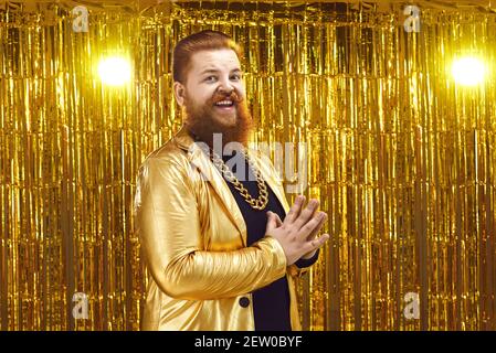 Divertente hipster uomo sovrappeso positivo in costume d'oro e catena in piedi con palme incrociate Foto Stock