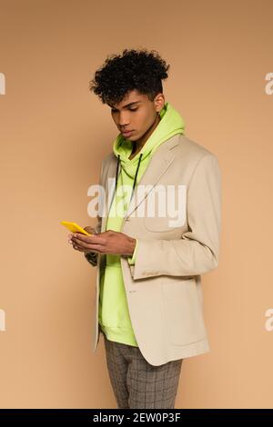 uomo afro-americano trendy in felpa con cappuccio e blazer con smartphone su beige Foto Stock