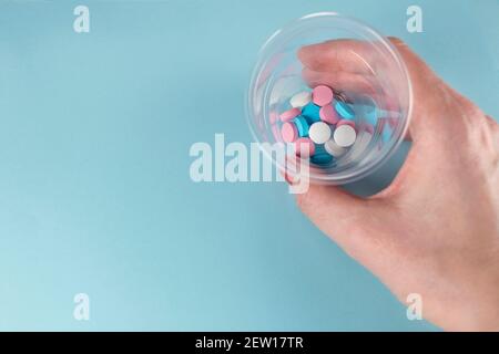 Le pillole di tenuta della mano femminile in una piccola tazza di medicina di plastica. Medicina e pillole. Farmaci multicolore su sfondo blu pastello. Foto Stock
