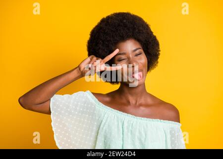 Ritratto fotografico di ragazza che mostra la lingua tenendo il segno V vicino all'occhio isolato su sfondo giallo brillante Foto Stock