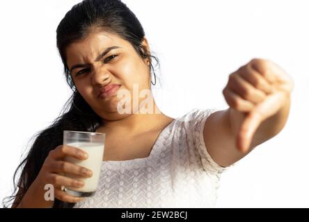 Una donna indiana / asiatica che tiene un bicchiere di latte con il volto infelice, guardando la macchina fotografica e pollici giù su sfondo bianco Foto Stock