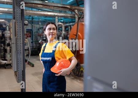 Il concetto di fabbrica industriale. Una giovane donna sorridente in uniforme tiene in mano un casco di sicurezza. Locale caldaia sullo sfondo. Foto Stock