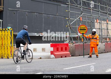 Un Marshall del traffico tiene in su il segno di arresto per arrestare una città solitaria del ciclista in arrivo di Londra, durante il lockdown Pandemic di Coronavirus Covid-19 Foto Stock