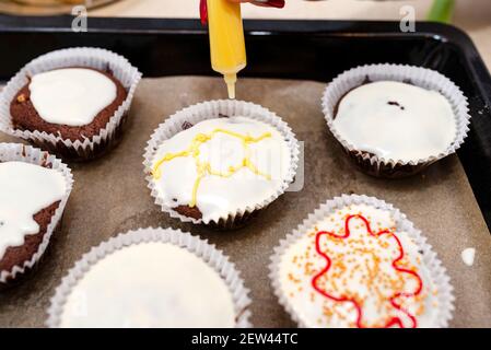 Una donna strizza la glassa colorata da un tubo su cupcake marrone cioccolato ricoperti di glassa bianca con decorazioni colorate. Foto Stock