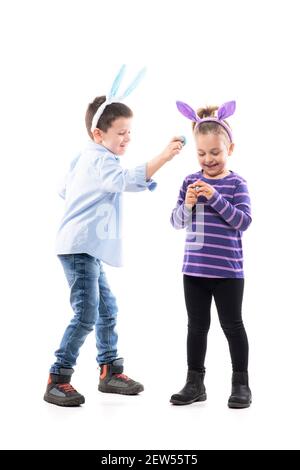 Fratello maggiore e sorella minore bambini con orecchie conigliate che giocano uova di Pasqua battendo o battendo gioco. Corpo intero isolato su sfondo bianco. Foto Stock