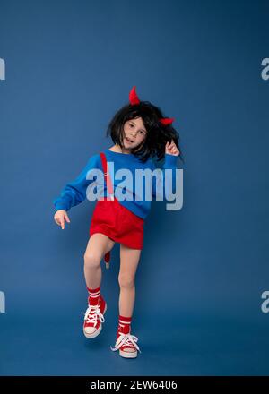 Bambina in parrucca imp con corna rosse indulgere in studio su sfondo blu. Concetto di costume di Carnevale Foto Stock