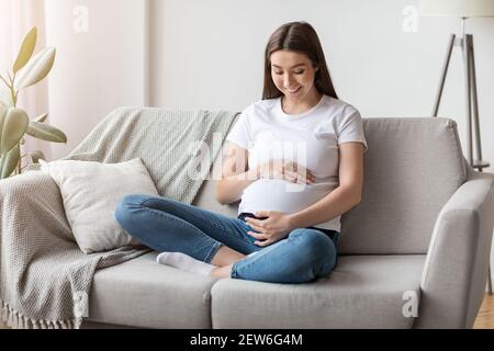 Bella giovane donna incinta abbracciando il pancino mentre si rilassa sul divano a casa Foto Stock