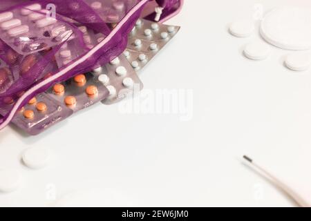 Varie pillole e un termometro elettronico fuoriuscito da una borsa in rete viola sul tavolo bianco. Farmaci per l'influenza, raffreddori. Pandemia di coronavirus. Io Foto Stock