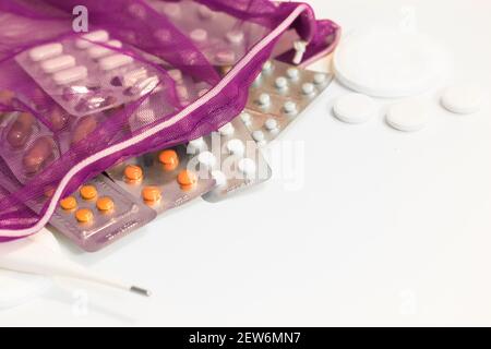 Varie pillole e un termometro elettronico fuoriuscito da una borsa in rete viola sul tavolo bianco. Farmaci per l'influenza, raffreddori. Pandemia di coronavirus. Io Foto Stock