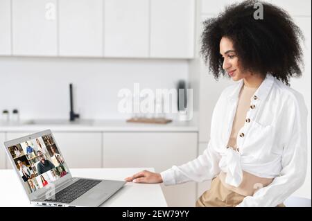 Freelance femminile afroamericana o titolare di affari con capelli ricci utilizzando l'app per la comunicazione video a distanza, lavorando o studiando online in remoto da casa, guardando un portatile con un gruppo di persone Foto Stock