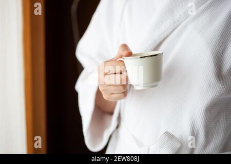 Un uomo in una vestaglia di terry bianca tiene una tazza di caffè davanti ad una finestra. Foto Stock
