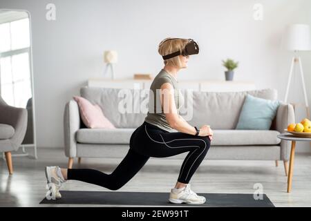 Allenamento virtuale. Vista laterale di una donna anziana in occhiali VR che fa affondo a casa. Sport intelligenti e tecnologie moderne Foto Stock