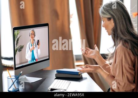 Online Medical Consultation.Grey-Haired donna anziana parla con un terapeuta medico femminile tramite videochiamata utilizza il computer, ottenendo consulenza medica, riceve risposte a domande e raccomandazioni Foto Stock