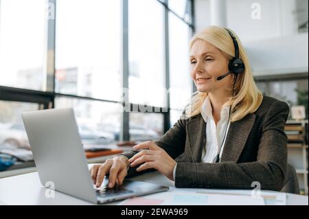 Una donna professionale matura e mirata utilizza il laptop, chattando con i colleghi. Elegante dipendente femminile in cuffia seduto alla scrivania, comunica con i colleghi tramite videochiamata Foto Stock