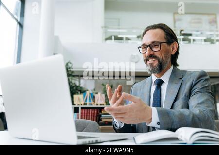 Un uomo d'affari maturo soddisfatto sta facendo una videochiamata con i colleghi, discutendo il progetto. Bel dipendente maschio usa il laptop, comunica con i colleghi, sorridendo Foto Stock
