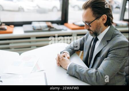 Dipendente maschile concentrato in occhiali sta imparando grafici finanziari, seduto alla scrivania. Serio uomo di mezza età che lavora in ufficio, sviluppando il programma di affari per l'azienda Foto Stock