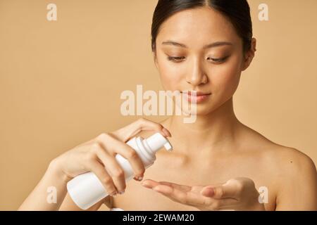 Studio ritratto di giovane donna naturale che tiene una bottiglia di schiuma delicata detergente viso isolato su sfondo beige. Prodotti di bellezza e concetto di cura della pelle Foto Stock