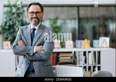 Ritratto di un convinto imprenditore di mezza età in abito elegante e occhiali in piedi in ufficio moderno, guardando la fotocamera e sorridente amichevole Foto Stock